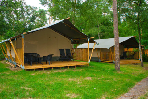 Tente safari avec toilette sur terrasse en bois, entourée d’herbe verte et d’arbres dans un camping paisible.
