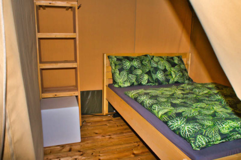 Gemütliches Bett mit grünem Bettzeug und Regal in einem Safarizelt bei Camping Aller-Leine-Tal.
