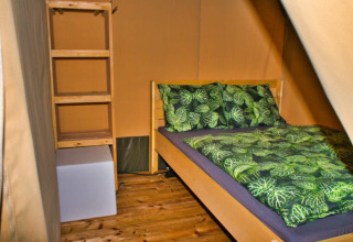 Gemütliches Bett mit grünem Bettzeug und Regal in einem Safarizelt bei Camping Aller-Leine-Tal.