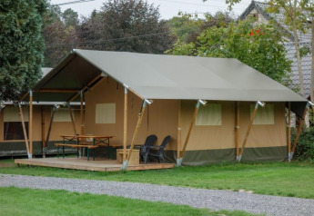 Photo of a large safari tent at a campsite with a covered deck, outdoor table, chairs and green lawn.