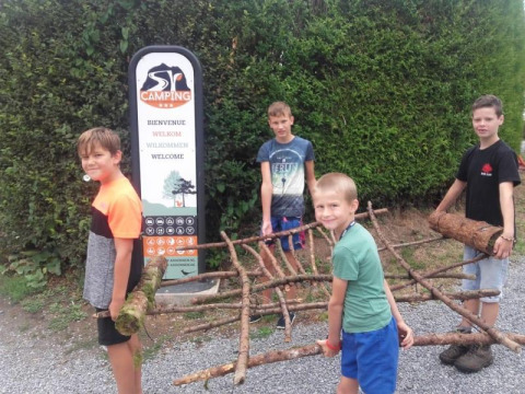 Quattro bambini trasportano rami davanti a un cartello di benvenuto alla Safari tent del Camping Village Sy in Belgio.