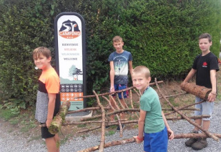 Quattro bambini trasportano rami davanti a un cartello di benvenuto alla Safari tent del Camping Village Sy in Belgio.