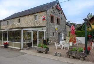 Edificio de piedra con terraza de cafetería en Camping Village Sy, Bélgica, con mesas, sillas y plantas.