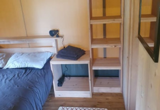 Binnenaanzicht van een safaritent met een enkel bed, houten rekken en een klein tapijt op houten vloer.