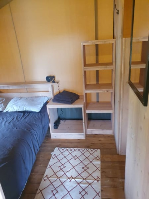 Binnenaanzicht van een safaritent met een enkel bed, houten rekken en een klein tapijt op houten vloer.