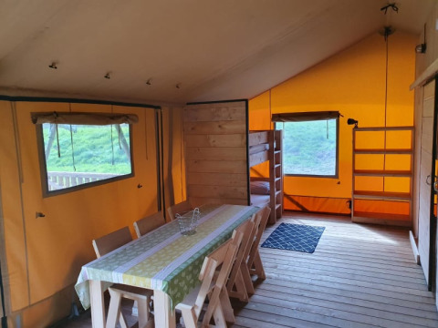 Vista interior de una tienda safari con mesa de comedor, sillas, litera y estanterías en Moulin du Pommier Camping.
