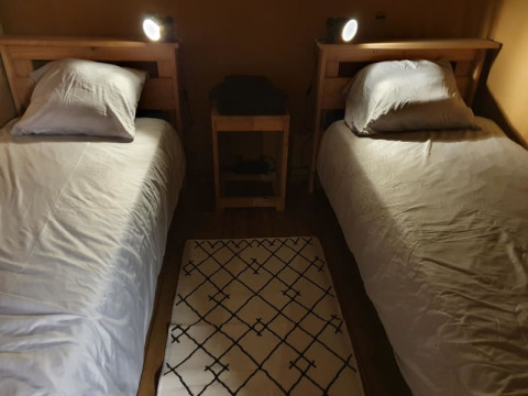 Dos camas individuales con sábanas blancas, cojines, mesita y alfombra sobre suelo de madera en tienda safari.