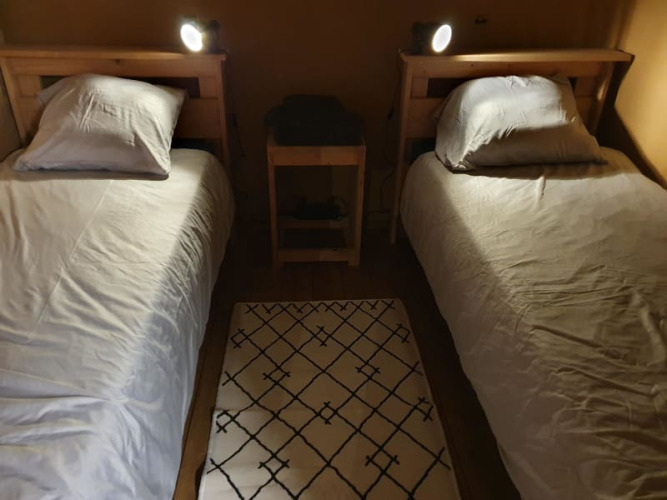 Zwei Einzelbetten mit weißen Bettlaken, Nachttisch dazwischen und Teppich auf Holzboden im Safarizelt.