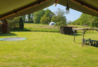Zicht vanuit safaritent op groen gras met ingegraven trampoline, houten hut en koepeltent achteraan.