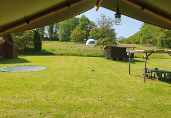 Blick aus Safari-Zelt auf grüne Wiese mit eingelassenem Trampolin, Holzschuppen und Kuppelzelt hinten.