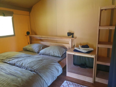 Safari tent interior at Moulin du Pommier Glamping & Camping, France, with double bed, wooden shelves and lamp.