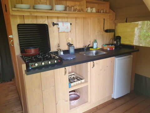 Pequeña cocina de camping con hornillo de gas, fregadero, cafetera, mini nevera y estantes de madera con platos.