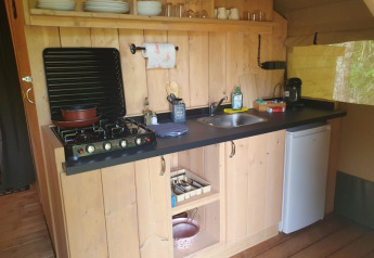 Pequeña cocina de camping con hornillo de gas, fregadero, cafetera, mini nevera y estantes de madera con platos.