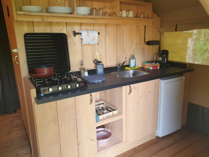 A small camping kitchen with gas stove, sink, coffee maker, mini fridge and wooden shelves with dishes.