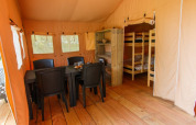 Interior of a safari tent with wooden floor, dining table, black chairs, shelves, and bunk beds visible.