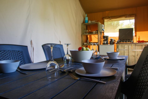 Dining table set in a safari tent with bowls, plates, wine glasses, and a small kitchen in the background.