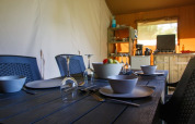 Dining table set in a safari tent with bowls, plates, wine glasses, and a small kitchen in the background.