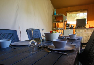 Dining table set in a safari tent with bowls, plates, wine glasses, and a small kitchen in the background.