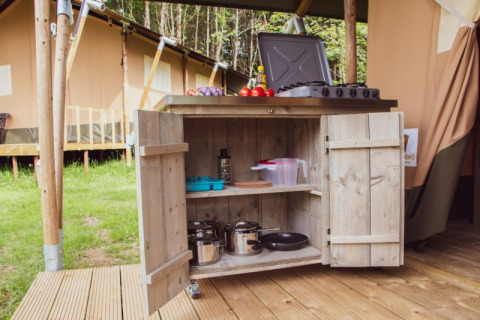 Cocina exterior con ollas y estufa frente a una tienda safari, rodeada de vegetación y árboles.