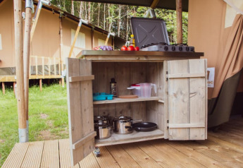 Cucina all'aperto con pentole e fornello davanti a una tenda safari, circondata dal verde e alberi.