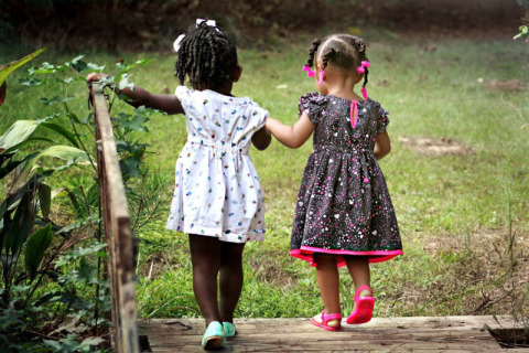 Dos niñas pequeñas con vestidos se dan la mano y caminan por un puente en el verde en Safari tent, Camping het Swinnenbos.