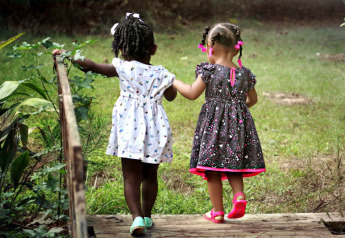 Dos niñas pequeñas con vestidos se dan la mano y caminan por un puente en el verde en Safari tent, Camping het Swinnenbos.