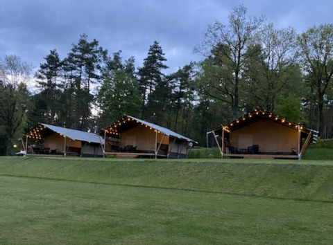 Safari tents with string lights at Camping Sonnenberg in Germany, set among trees and green lawn at dusk.
