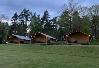 Safari tents with string lights at Camping Sonnenberg in Germany, set among trees and green lawn at dusk.