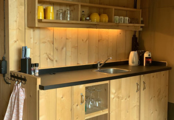 A cozy wooden kitchenette in a safari tent, featuring a black countertop, sink, and open shelving.