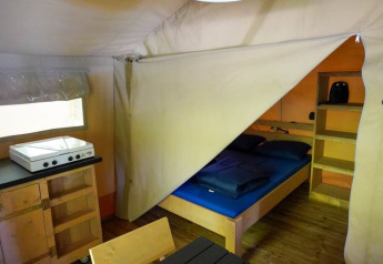 Vista interior de una tienda safari en Camping Pittoresque, Francia, con cama, estantes y cocina auxiliar.