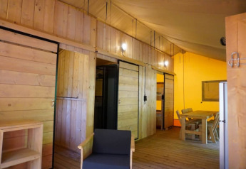 Vista interior de una tienda safari con paredes de madera, mesa de comedor, sillas y zona de dormir en Camping Pittoresque.
