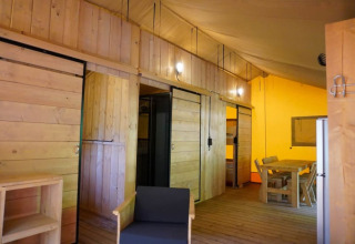 Binnenzicht van een safaritent met houten wanden, eettafel, stoelen en slaapgedeelte op Camping Pittoresque.