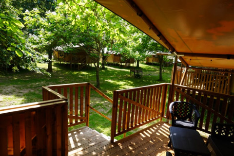 Holzterrasse eines Safarizelts mit Bad auf Camping Pittoresque in Frankreich, umgeben von Bäumen.