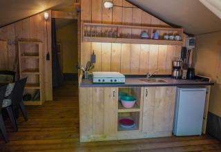 Gezellige safaritent keuken met houten panelen, kookplaat, spoelbak en koelkast bij Camping Pittoresque in Frankrijk.