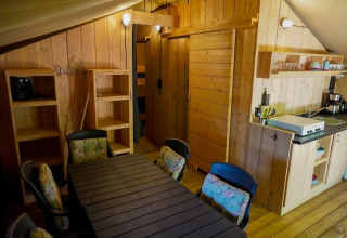 Indvendigt billede af et safari telt med træinteriør, spisebord, køkkenområde og reol på Camping Pittoresque.