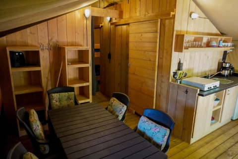 Indvendigt billede af et safari telt med træinteriør, spisebord, køkkenområde og reol på Camping Pittoresque.