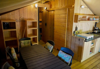 Interior view of a safari tent with wooden walls, dining table, kitchen area, and shelves at Camping Pittoresque.