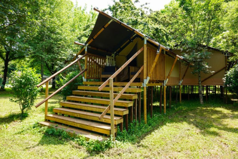 Photo of a safari tent named Duo Lodge, elevated on wooden stilts, surrounded by lush green trees and grass.