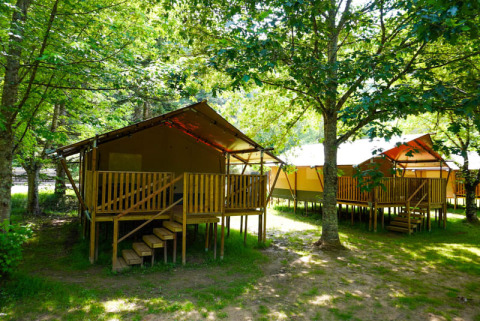 Safari tent met toilet, verhoogd op houten platform, gelegen tussen bomen in een groene campingomgeving.