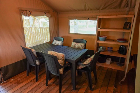 Zona pranzo in tenda safari con pavimento in legno, tavolo, sedie e scaffali al Camping Pittoresque, Francia.