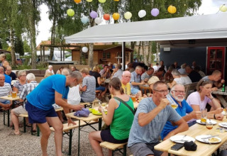 Menschen essen und trinken an langen Tischen unter Lampions beim Safarizelt auf Camping Elbeling in Deutschland.