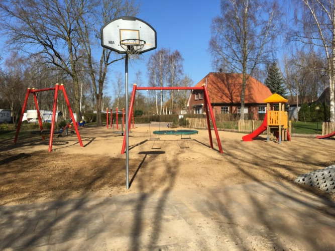 Playground with swings, slide and basketball hoop at Camping Elbeling under a clear blue sky in Germany.