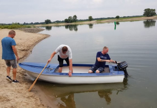 Three men launch a small boat into the water near a safari tent, by a tranquil lakeshore setting.