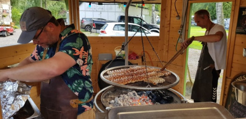 Zwei Männer grillen Würste über Holzkohle in einer offenen Küche eines Safarizelts mit Holzwänden.