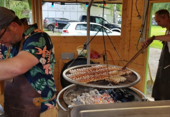 Zwei Männer grillen Würste über Holzkohle in einer offenen Küche eines Safarizelts mit Holzwänden.