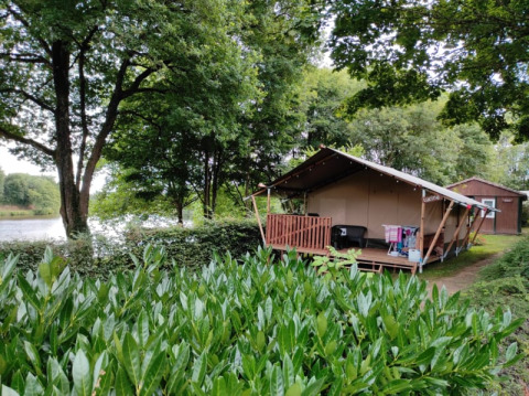 Tenda safari con veranda in legno immersa nel verde, situata accanto a un lago con un'altra cabina sullo sfondo.
