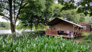 Safari tent with wooden porch set among lush greenery beside a lake, with another cabin in the background.