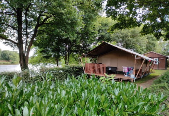 Tienda safari con porche de madera entre vegetación exuberante junto a un lago y una cabaña al fondo.