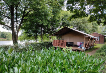 Tenda safari con veranda in legno immersa nel verde, situata accanto a un lago con un'altra cabina sullo sfondo.