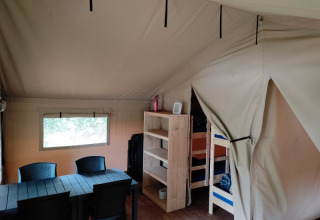 Interior de la tienda safari en Camping L’Etruyère, Francia, con mesa, sillas, estantería y literas.